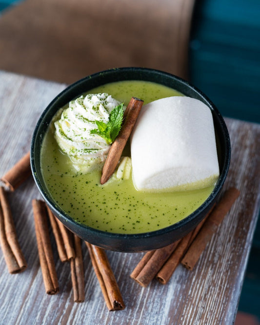 Hot white chocolate with matcha served in a bowl with marshmallow and whipped cream, surrounded by cinnamon sticks